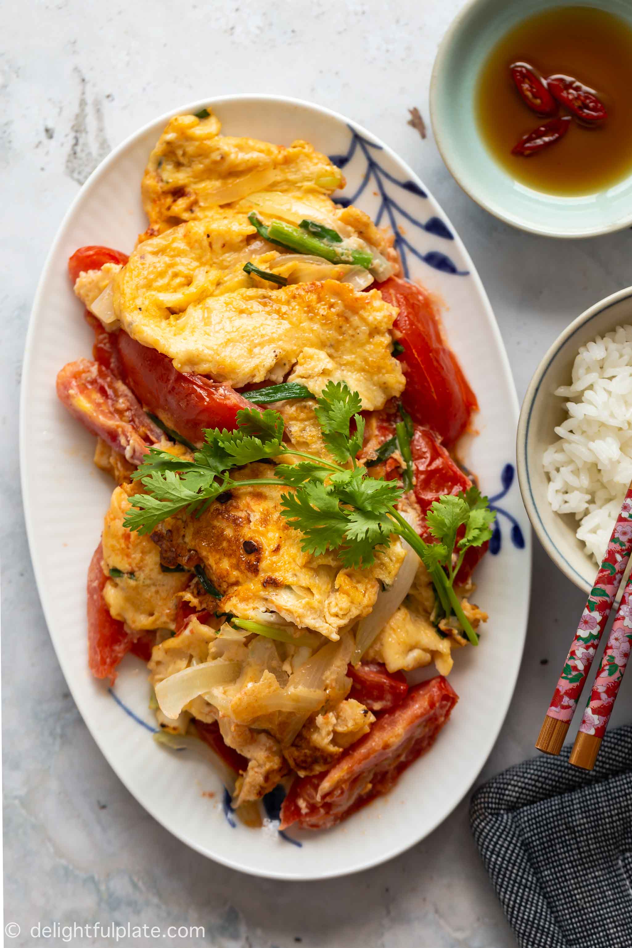 a serving plate with pan fried eggs with tomatoes and onions, decorated with cilantro and served with rice and fish sauce on the side